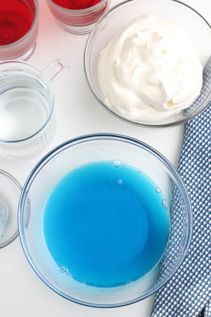A bowl of blue liquid gelatin, a bowl of whipped topping, and a measuring cup on a white surface with a blue towel.