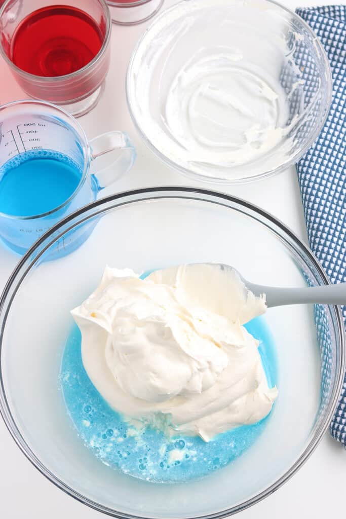 A mixing bowl with blue gelatin and whipped topping, surrounded by red and blue liquids and an empty bowl.