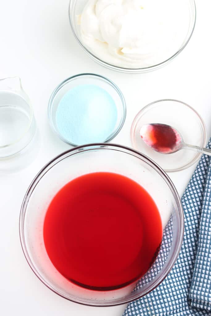 A bowl of dissolved red gelatin, a bowl of whipped cream, blue powdered jello, water, and a spoon on a checkered cloth.