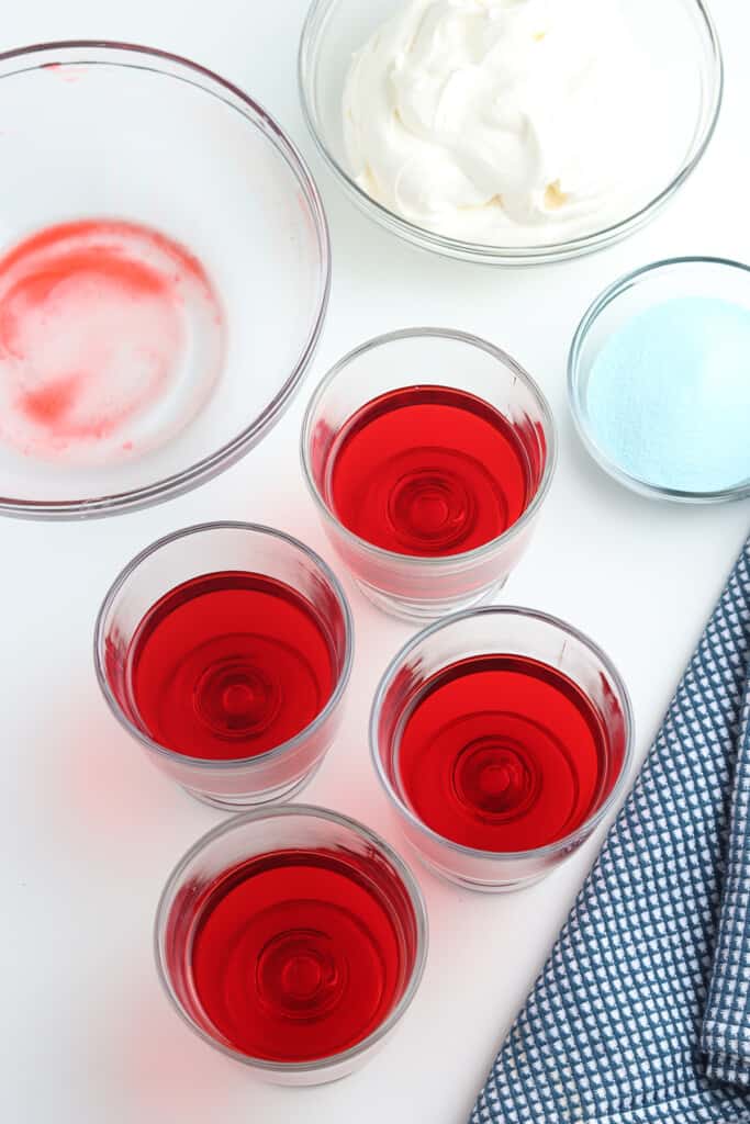 Four glasses of red gelatin, a bowl of whipped cream, and a blue checkered cloth on a white surface.