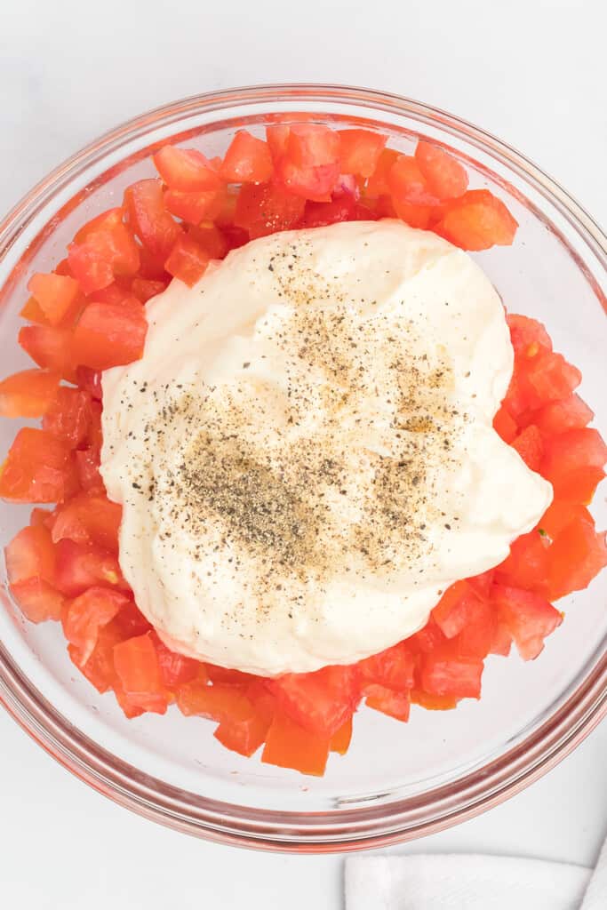 Diced tomatoes topped with mayonnaise and black pepper in a clear glass bowl on a white surface.
