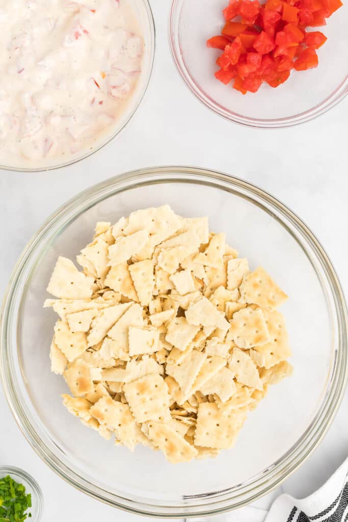 Glass bowls with crushed crackers, diced tomatoes, creamy mixture, and chopped green herbs on a white surface.