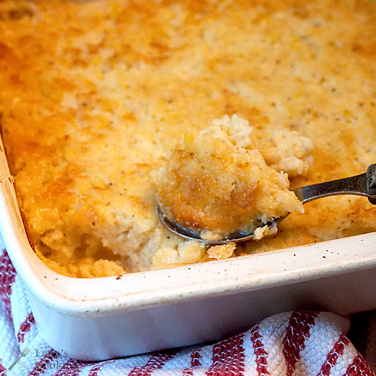 A metal spoon scoops a portion of a baked, golden-brown corn casserole from a white rectangular baking dish, which rests on a red and white striped cloth.