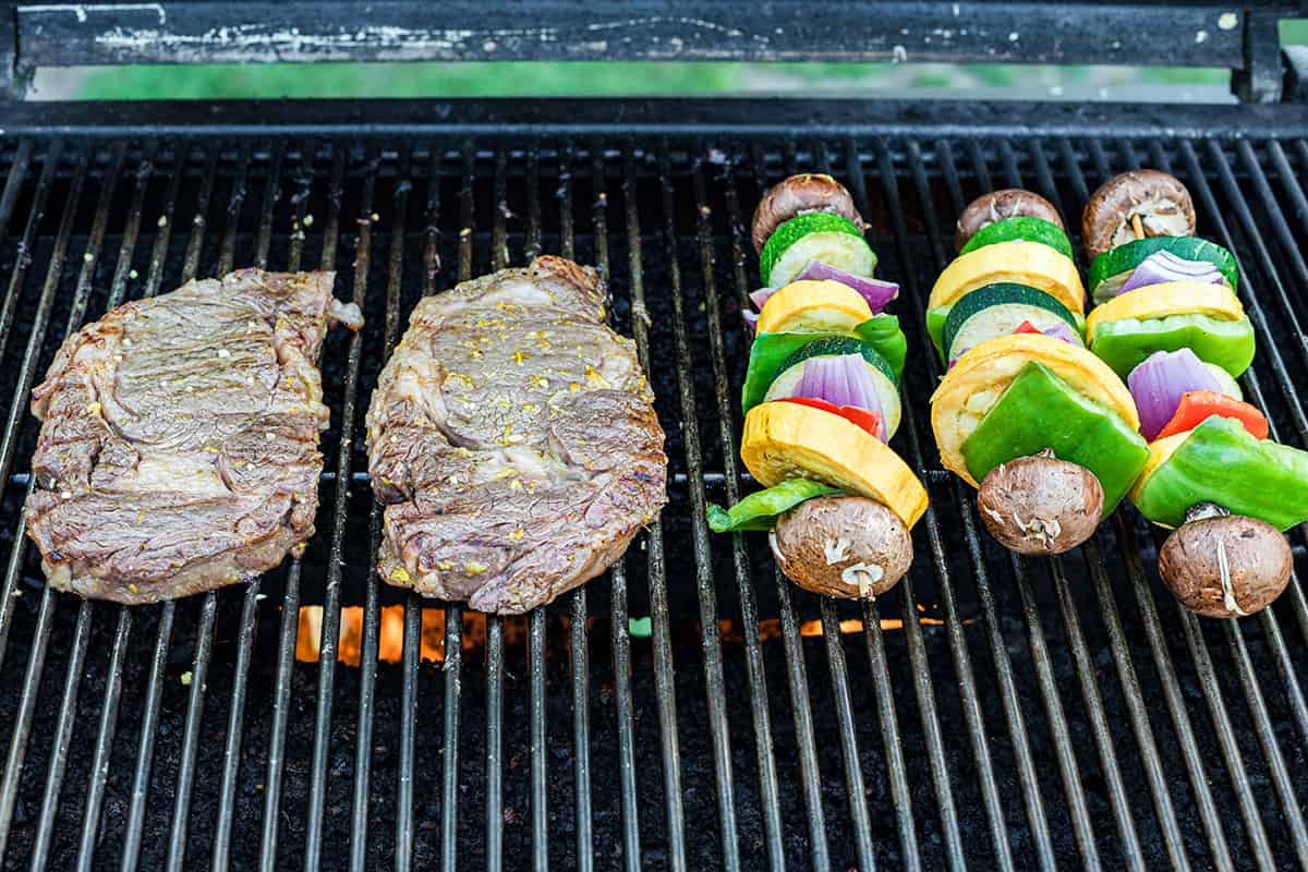 Steaks and vegetable skewers cooking on a grill.