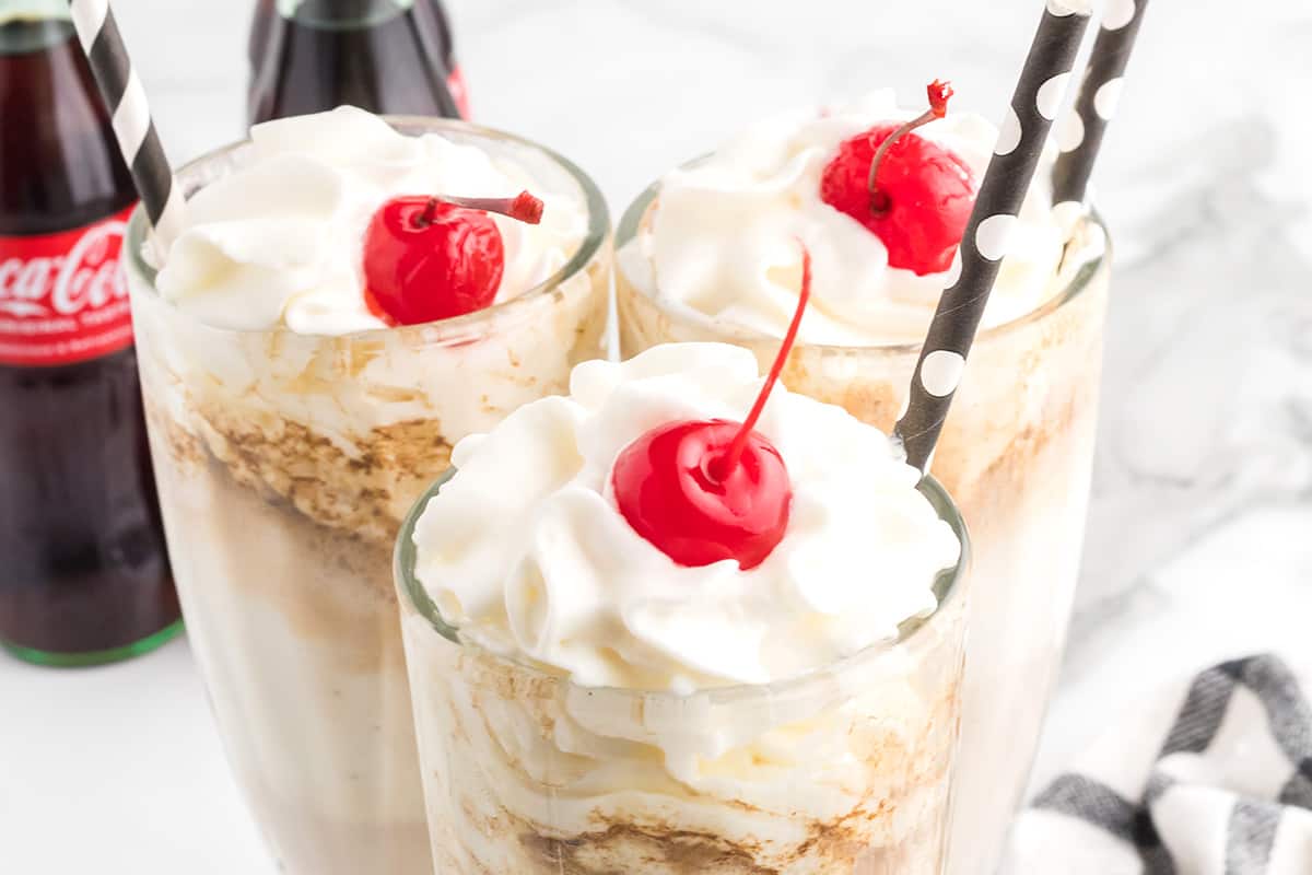 Coke floats in tall glasses with whipped cream and a cherry on top.