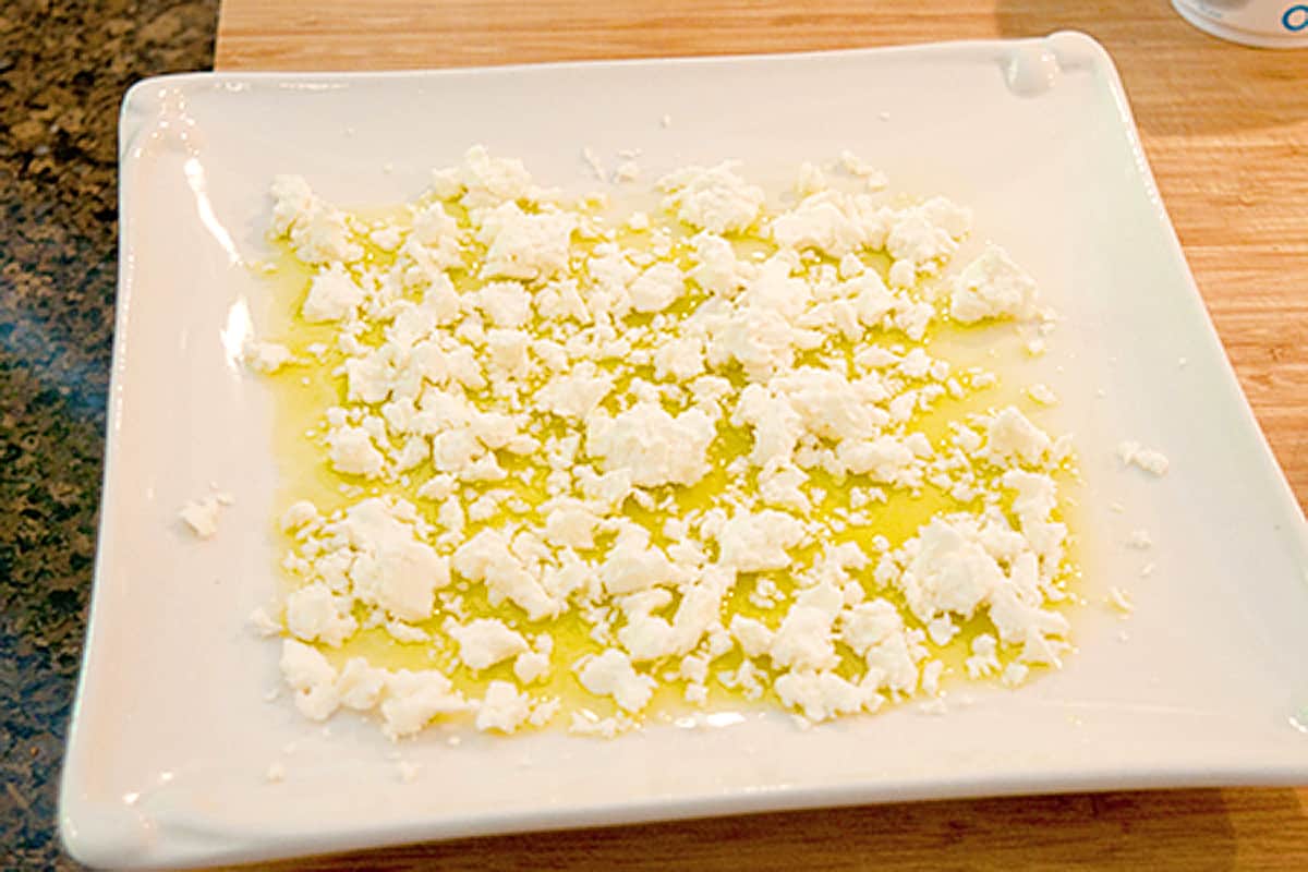 Crumbled feta cheese added to olive oil on a white platter.