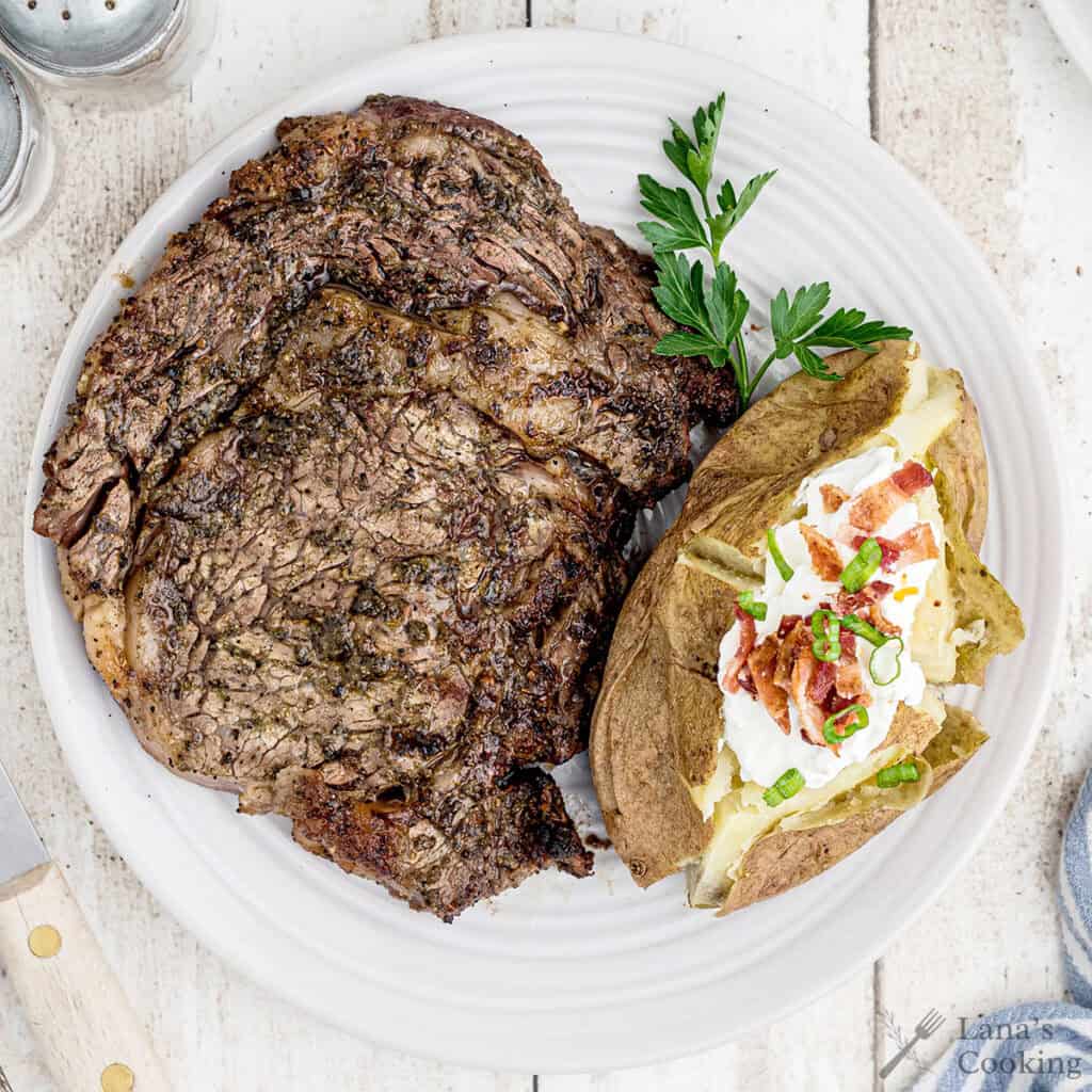 A grilled steak and a baked potato topped with sour cream, bacon, and green onions are served on a white plate, garnished with a sprig of parsley.