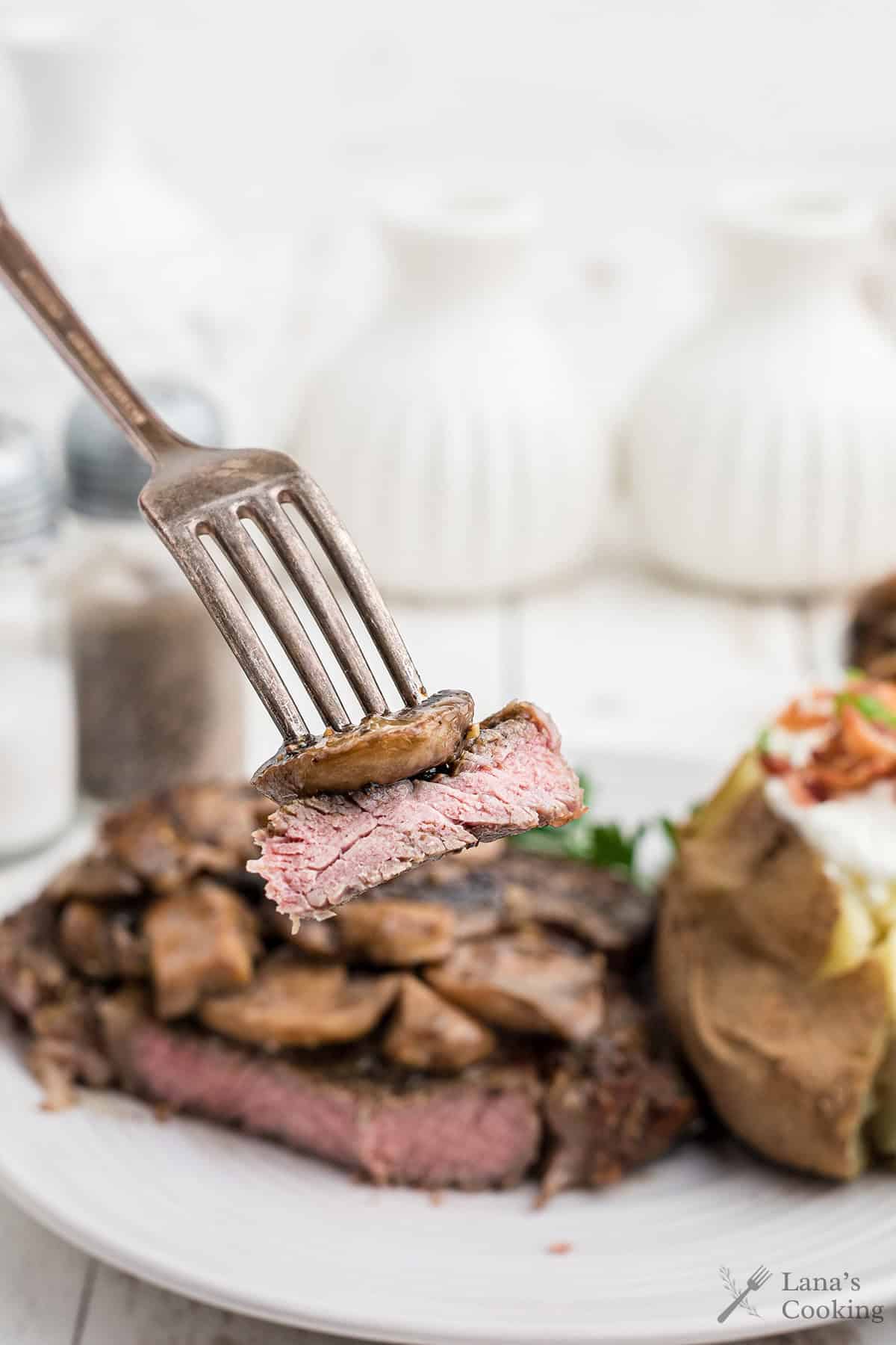 A fork holds a bite of steak topped with sautéed mushrooms above a plate with more steak and a baked potato in the background.