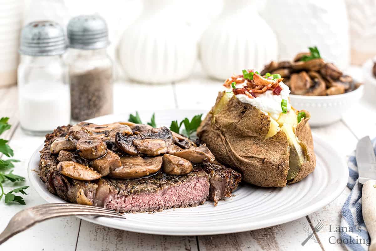 A plate with a grilled steak topped with sautéed mushrooms and a baked potato with sour cream, bacon, and chives. Salt and pepper shakers are in the background.
