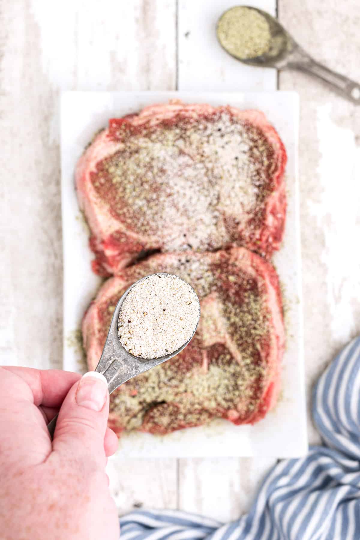 Sprinkling seasonings on two ribeye steaks.