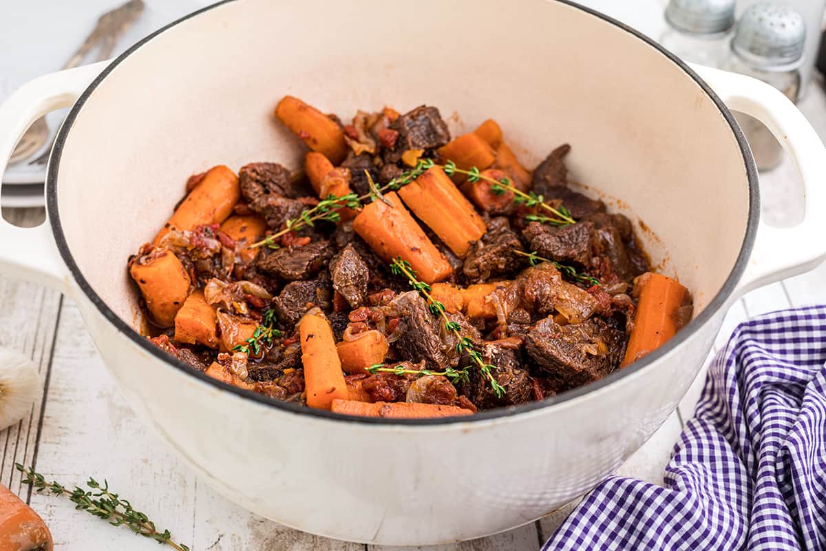 Beef stew with carrots and herbs in a white pot on a wooden table, next to a checkered cloth.