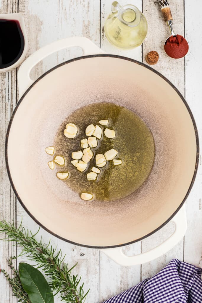 Garlic cooking in olive oil in a large white pot on a rustic wooden surface with herbs nearby.