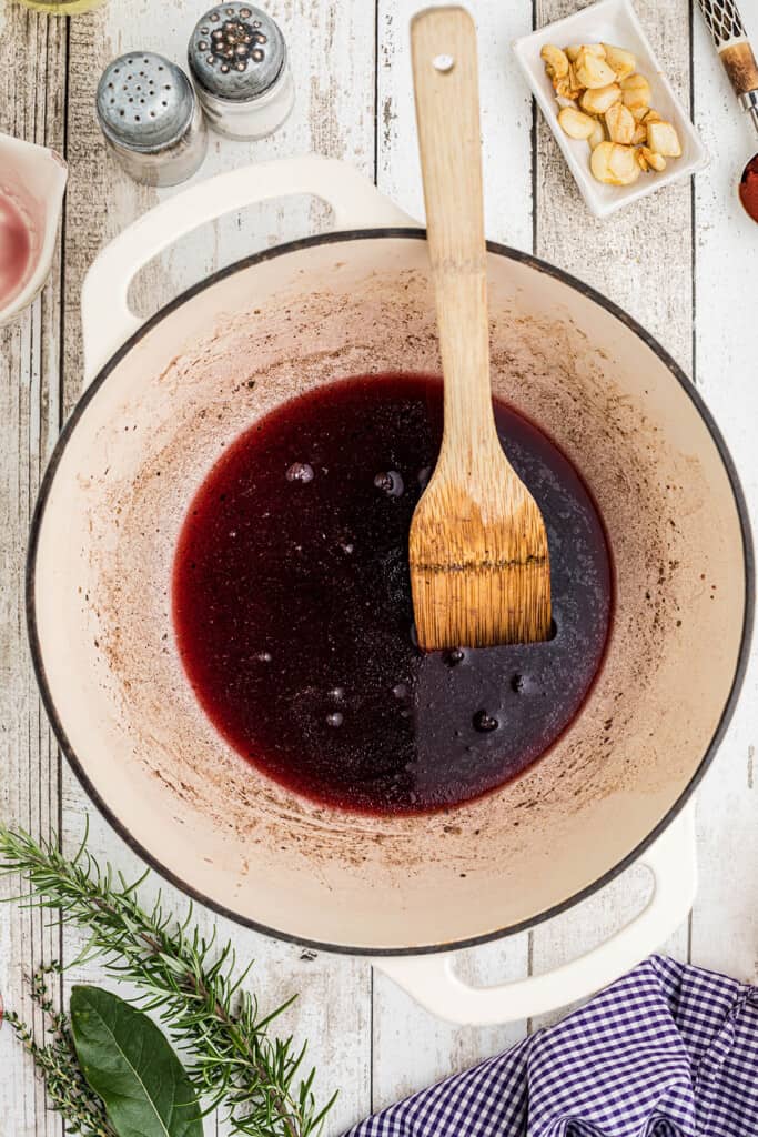A pot with red sauce, a wooden brush, herbs, garlic, and a towel on a rustic wooden table.