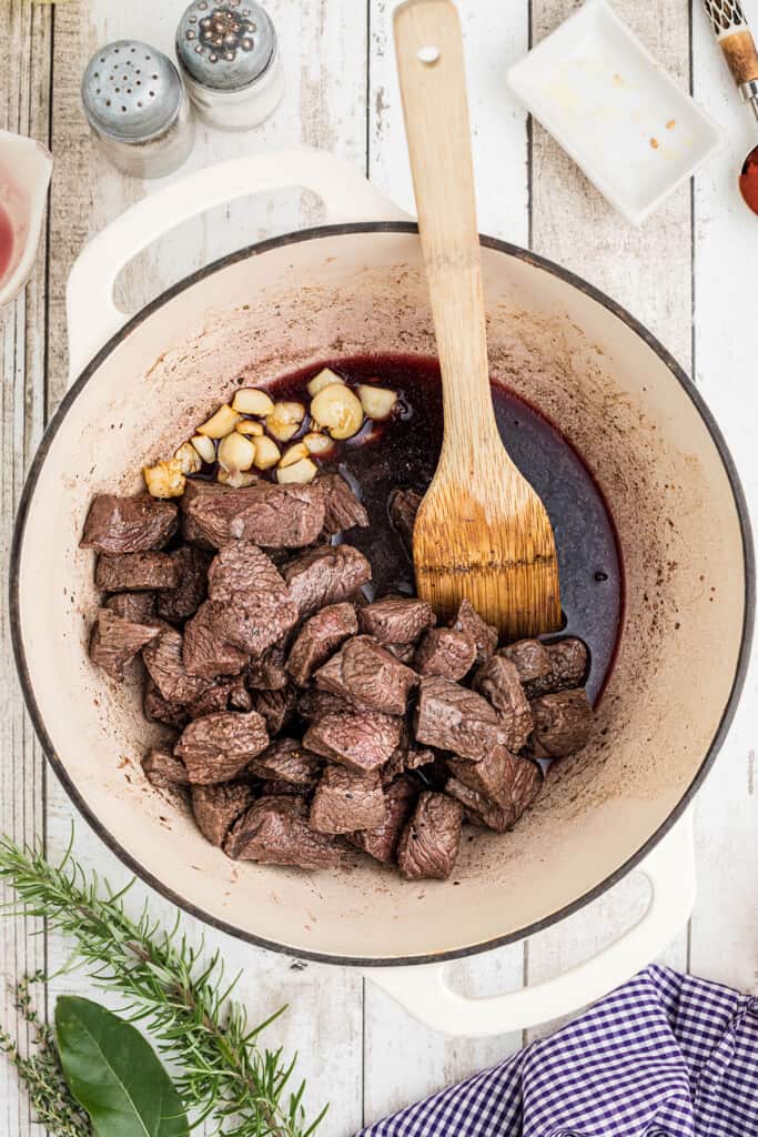 Chunks of beef, garlic cloves, and red wine simmer in a Dutch oven with a wooden spoon.