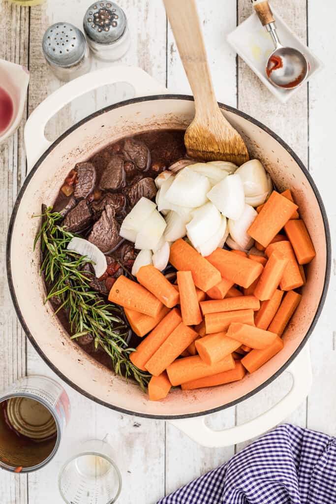Chopped carrots, onions, herbs, and beef in a pot with a wooden spoon, ready to cook.
