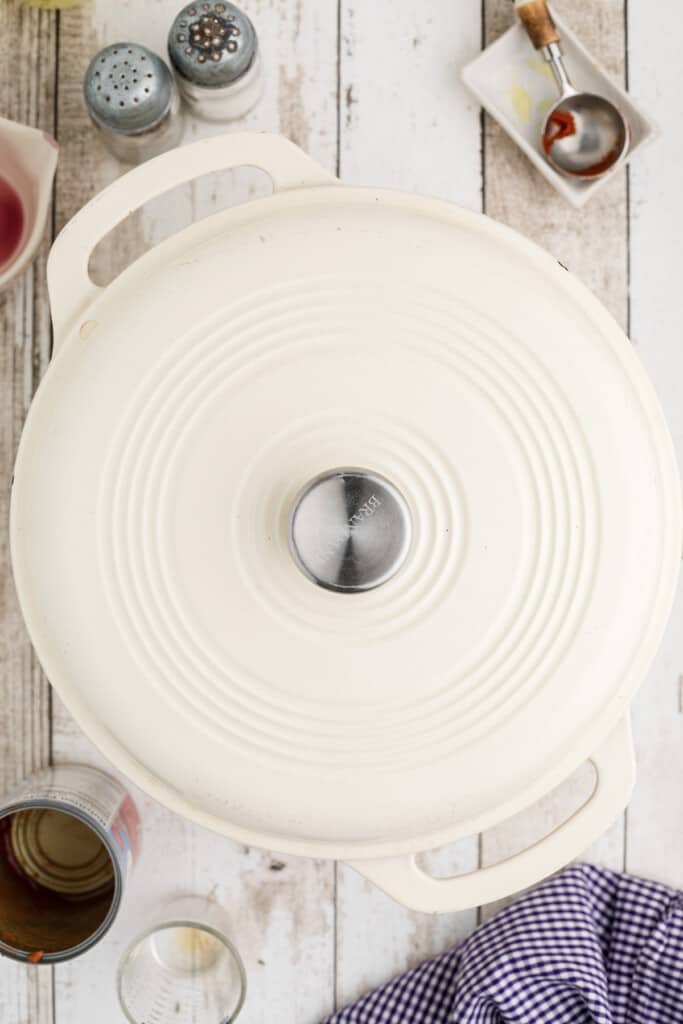 A white Dutch oven with a lid sits on a rustic wooden table next to kitchen items and a blue checkered cloth.