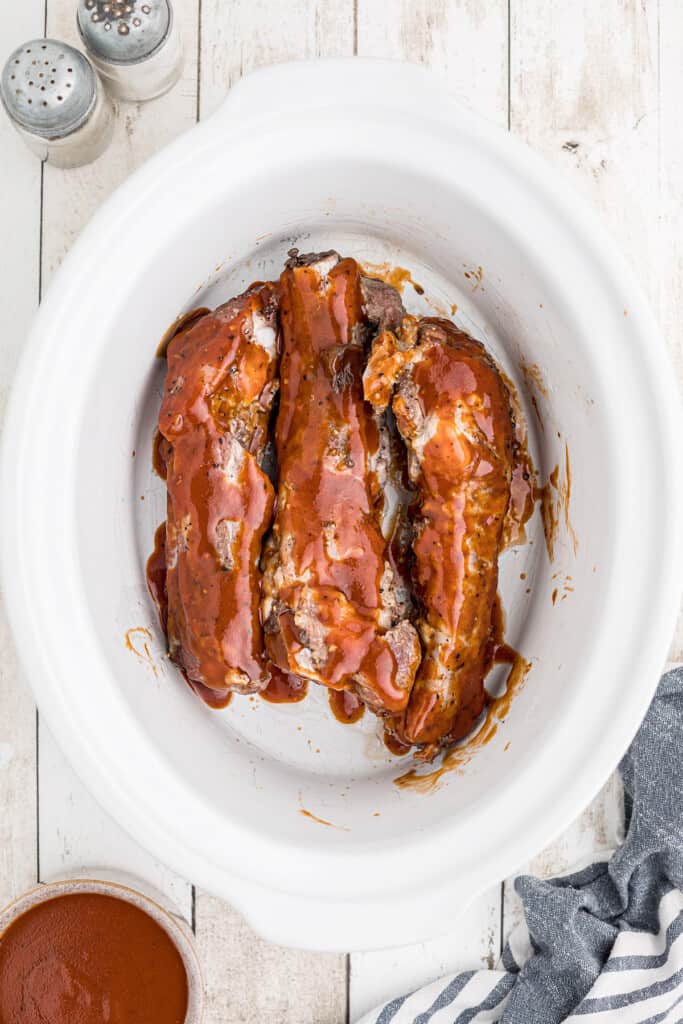 Spareribs covered in barbecue sauce in a white slow cooker, with salt and pepper shakers nearby.