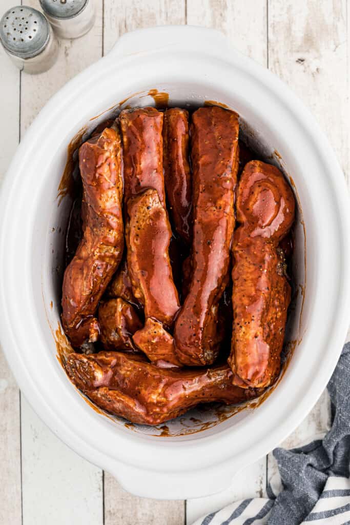 Pork ribs coated in barbecue sauce arranged in a white slow cooker, ready for cooking.