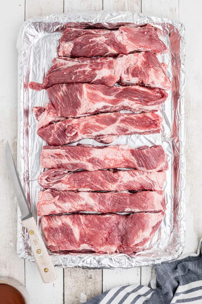 Raw pork ribs arranged on a foil-lined baking sheet with a knife placed beside them on a white wooden surface.