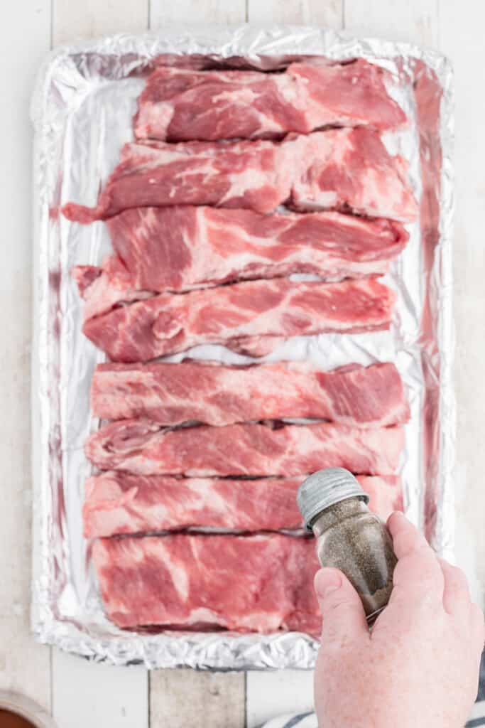 A hand sprinkling seasoning over raw pork ribs on a foil-lined baking sheet.