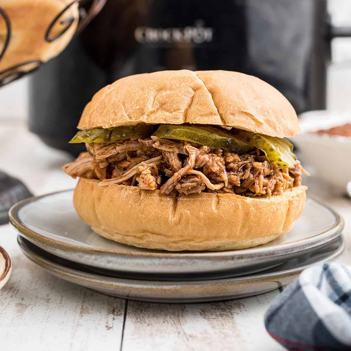 A pulled pork sandwich on a serving plate with a slow cooker in the background.