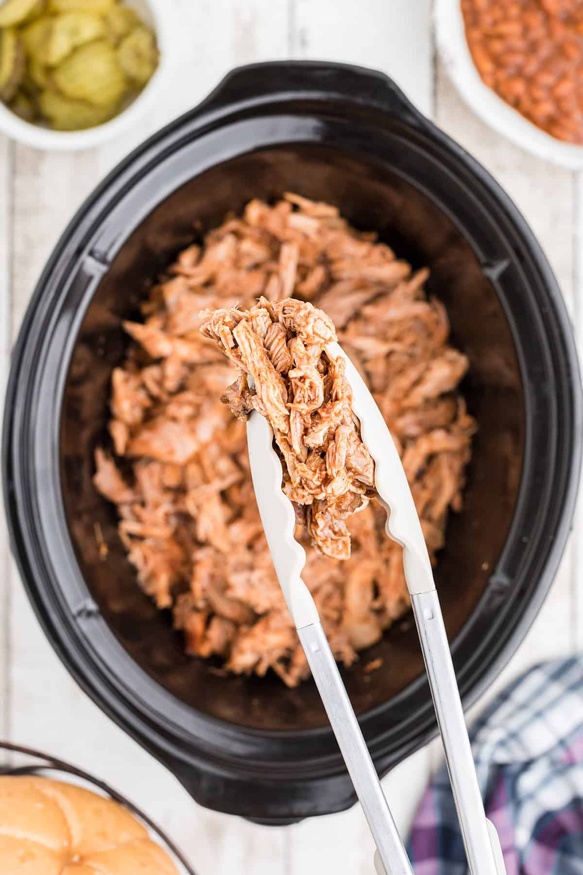 Tongs holding a portion of pulled pork above a slow cooker.