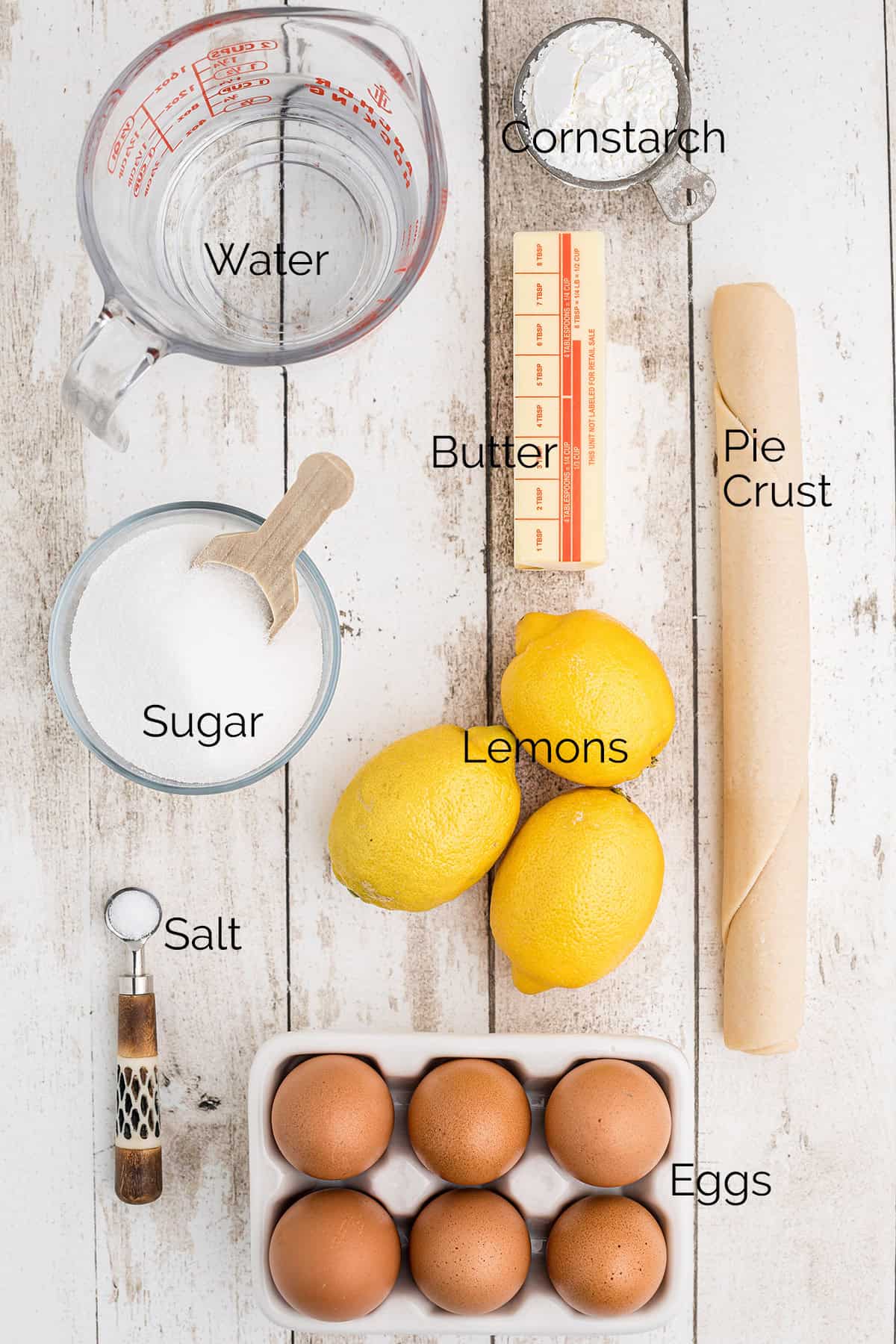 Top-down view of labeled lemon pie ingredients: water, cornstarch, butter, pie crust, sugar, lemons, salt, and eggs.