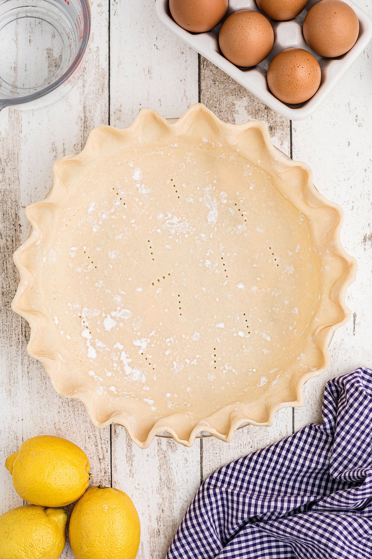 Unbaked pie crust in a fluted pan, surrounded by eggs, lemons, a measuring cup, and a checkered kitchen towel.