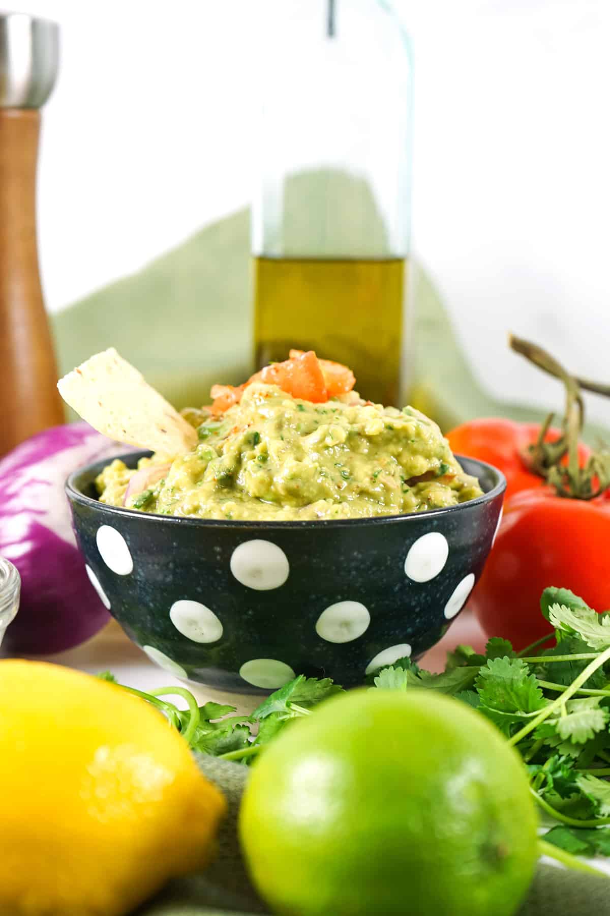 Finished guacamole in a decorative bowl surrounded by ingredients used in the recipe.