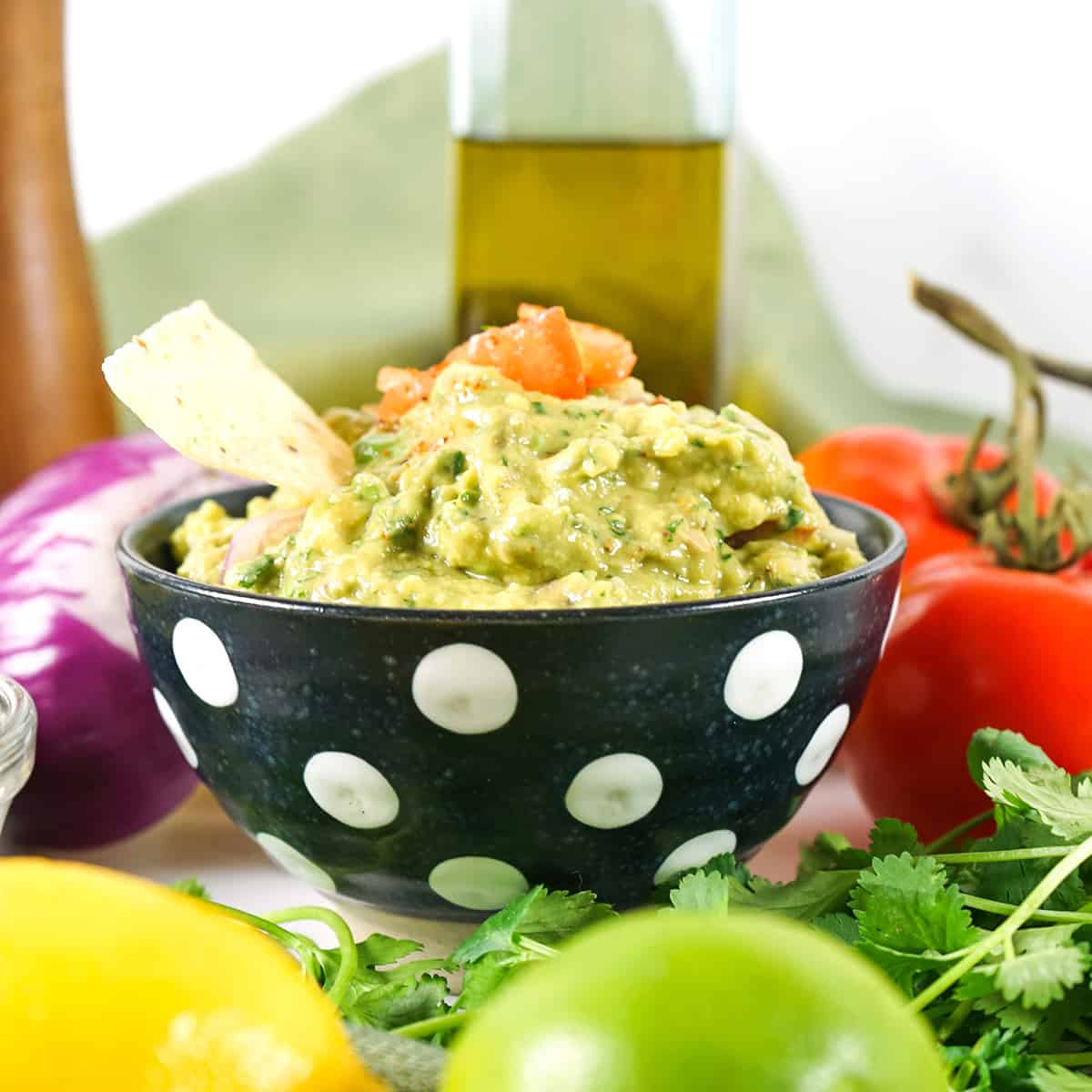 Finished guacamole in a decorative bowl surrounded by ingredients used in the recipe.
