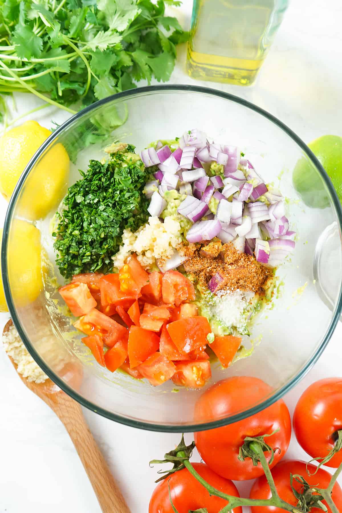 Glass bowl with chopped tomatoes, red onion, herbs, and spices, surrounded by lemons, cilantro, and tomatoes.