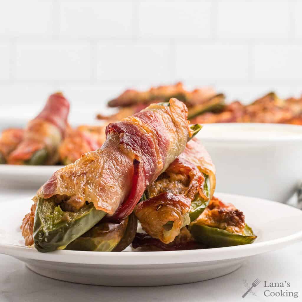 Bacon-wrapped stuffed jalapeños stacked on a white plate with more in the background.