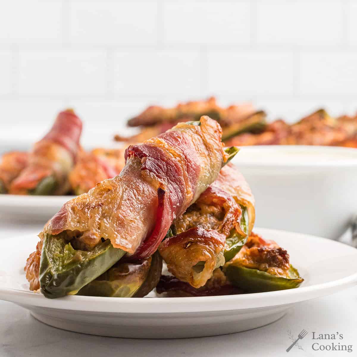 Bacon-wrapped stuffed jalapeños stacked on a white plate with more in the background.