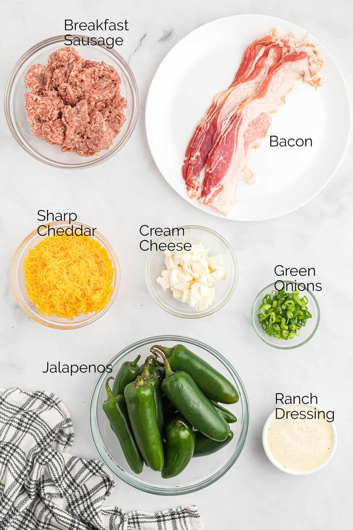 Bowls of breakfast sausage, bacon, cheddar, cream cheese, green onions, jalapenos, and ranch on a white surface.
