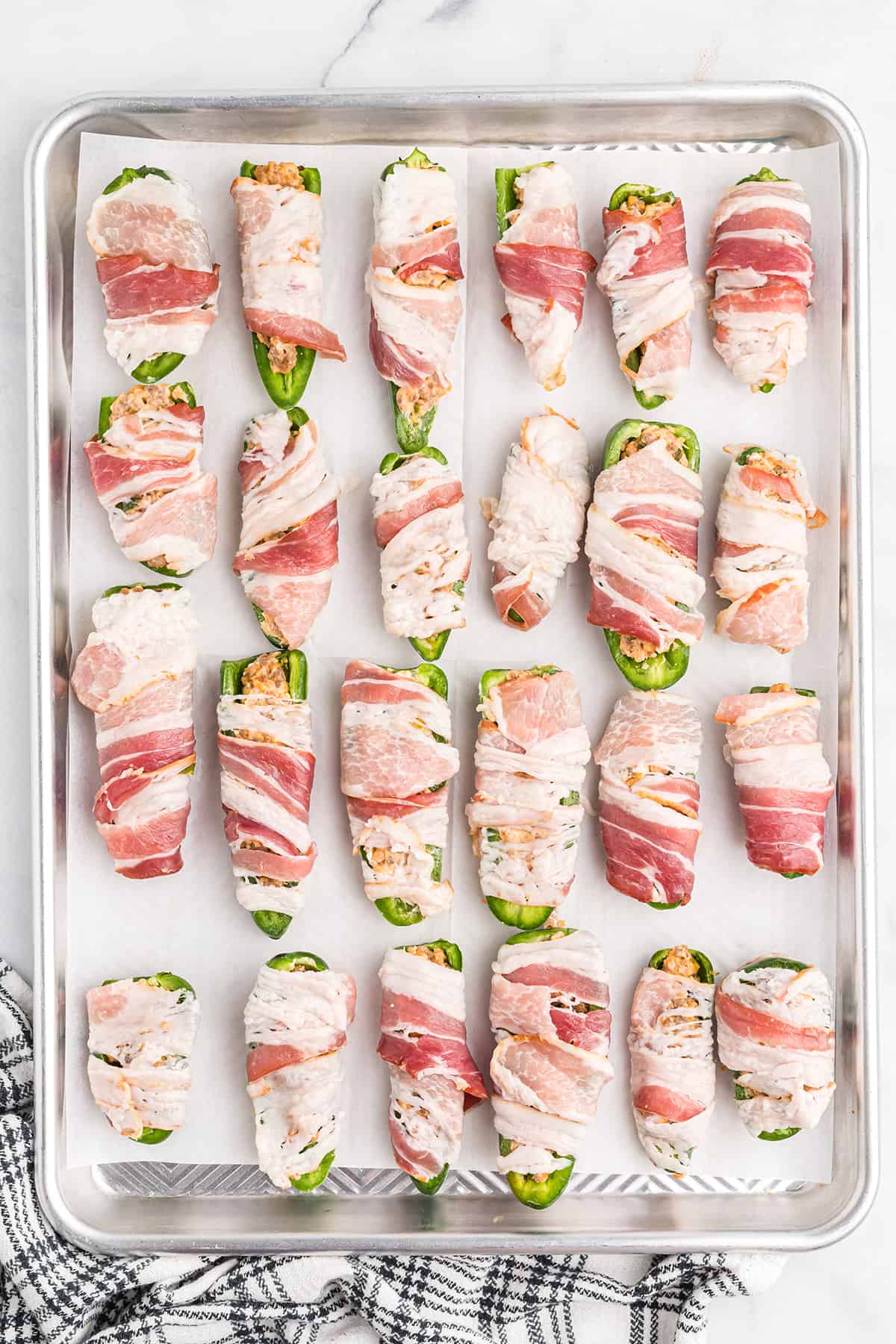 Jalapenos filled and wrapped with bacon on a baking sheet.
