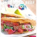 Two beef tacos with tomatoes, onions, and cilantro on a plate, with lime wedges in the background.