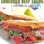 Two tacos filled with shredded beef, diced tomatoes, sliced red onions, and fresh cilantro are on a white plate. A lime wedge sits in the background. The image features the text Slow Cooker Shredded Beef Tacos.