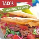 Two tacos filled with shredded beef, diced tomatoes, pickled red onions, and cilantro are shown on a plate. Lime wedges are visible in the background. Text on the image reads “Slow Cooker Shredded Beef Tacos with Pickled Red Onions.”.
