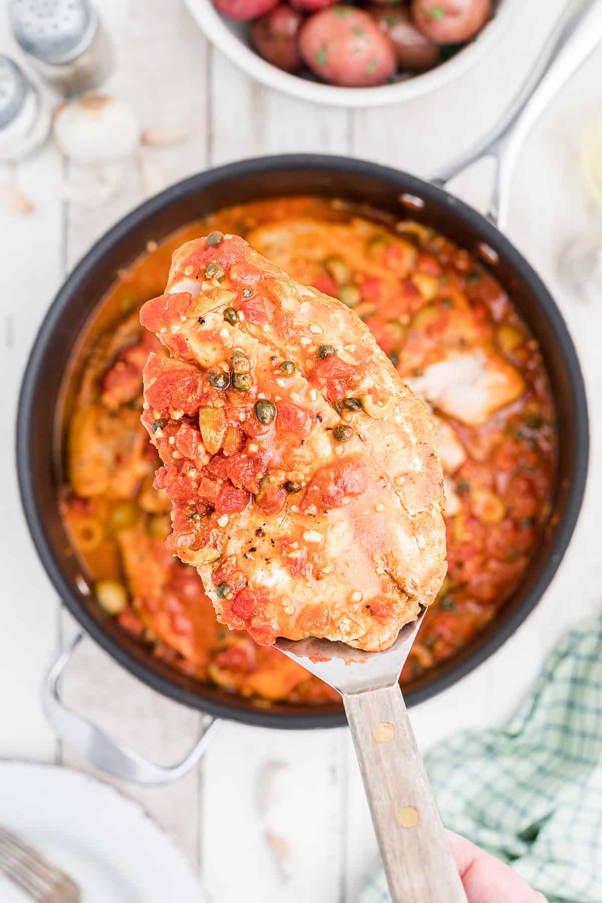 A spatula holds a piece of fish topped with chunky tomato sauce over a skillet of the same dish.