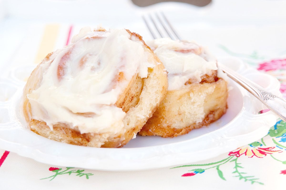 Two frosted cinnamon rolls on a white plate with a fork, set on a floral-patterned tablecloth.