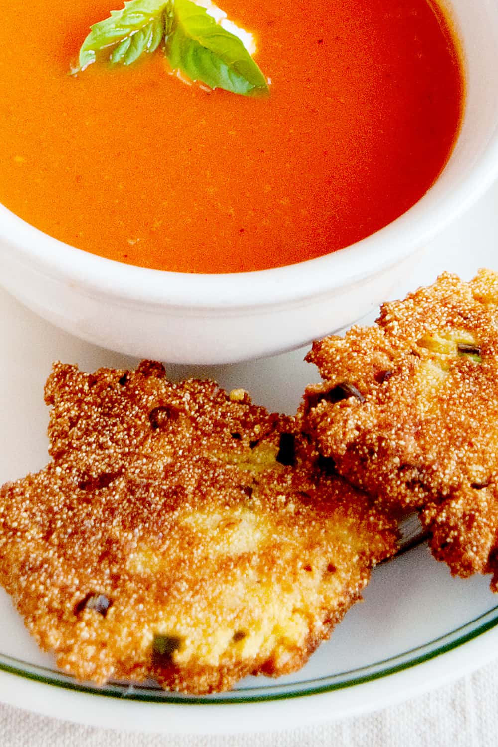 Two cornmeal scallion fritters on a plate alongside tomato soup.