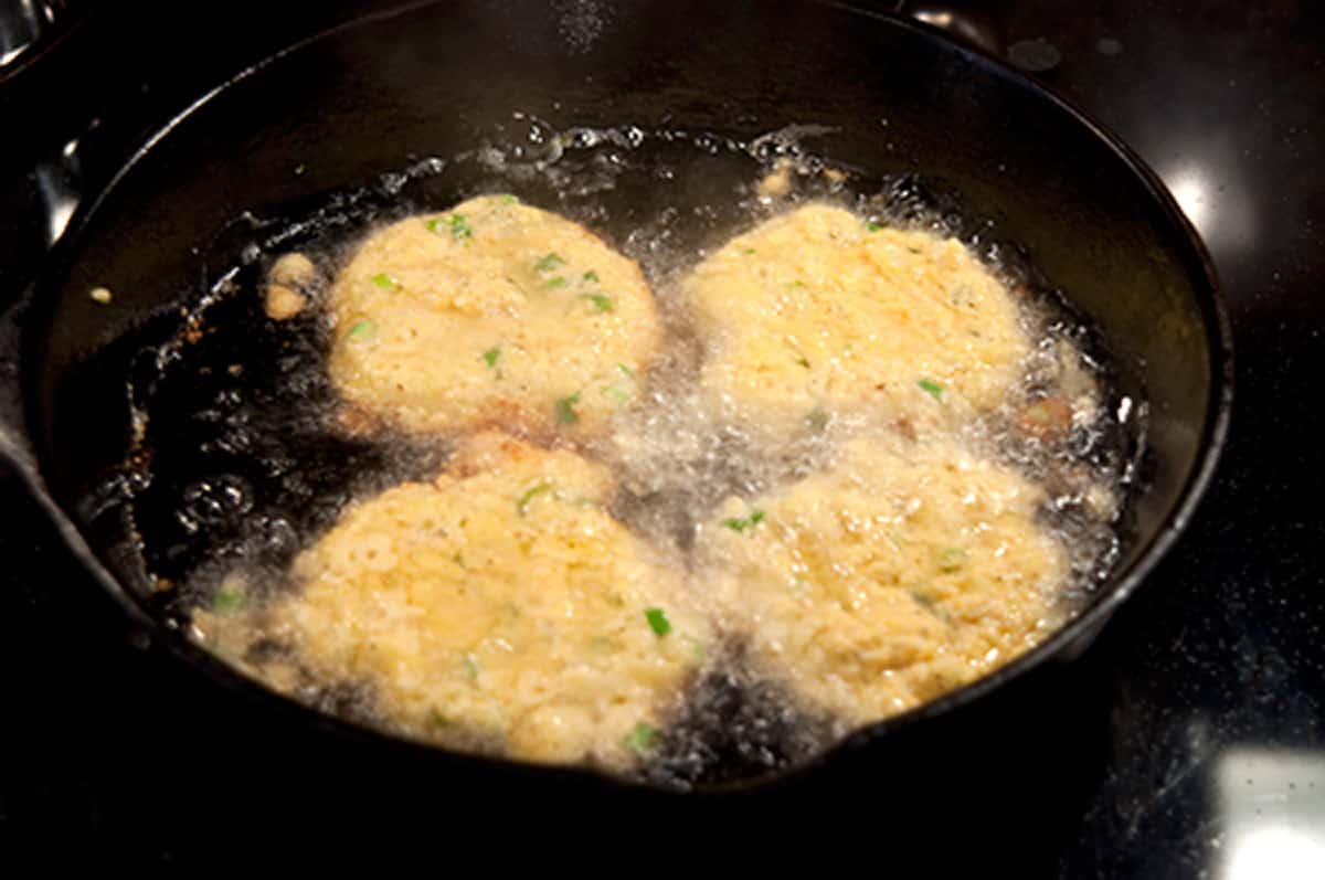 Fritters frying in hot oil in a black iron skillet.