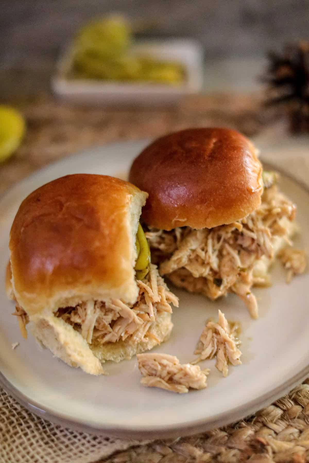 Two shredded chicken sliders on soft buns, served on a plate with pickles in the background.