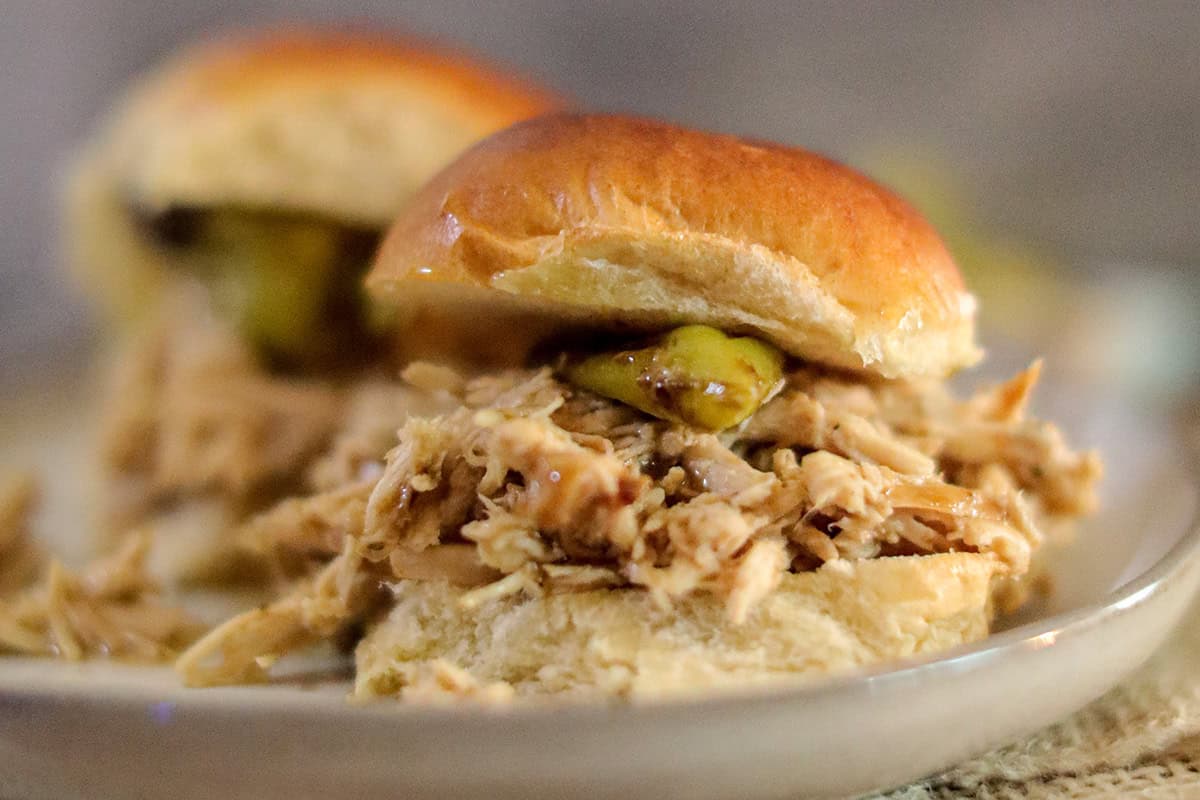 Pulled chicken slider with a pickle slice on a soft bun, served on a plate.