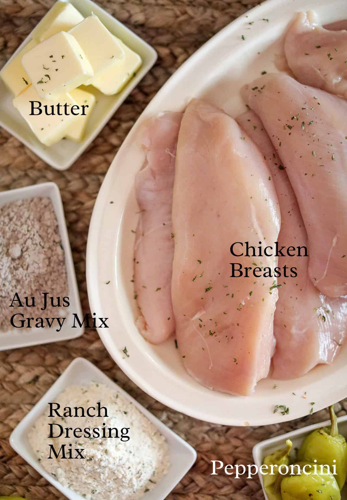 Raw chicken breasts on a plate with butter, au jus gravy mix, ranch dressing mix, and pepperoncini on the side.