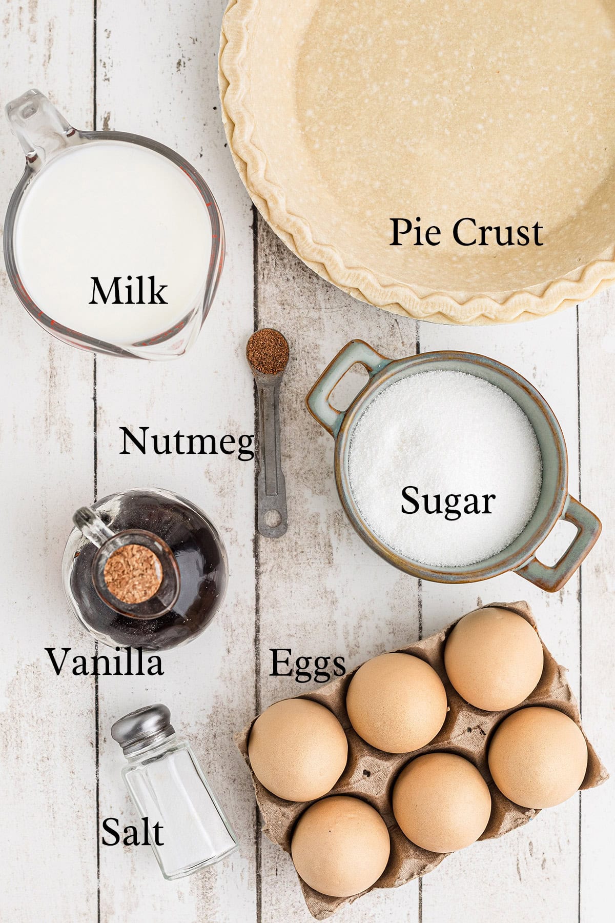 Ingredients for a pie: pie crust, milk, nutmeg, sugar, vanilla, eggs, and salt arranged on a table.
