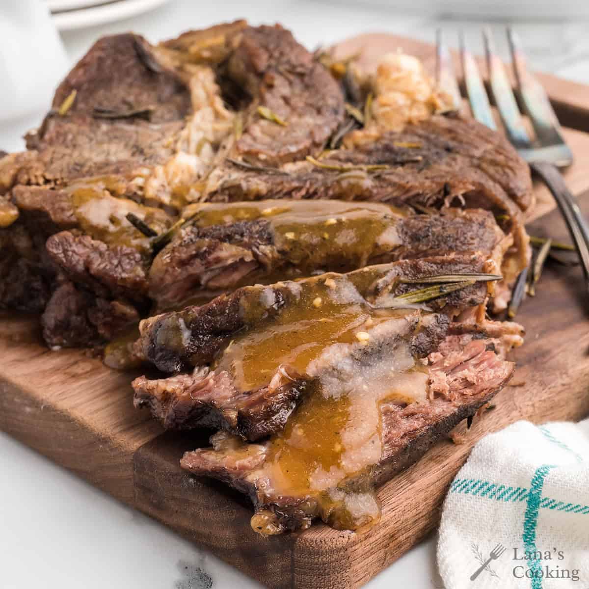 Sliced pot roast with gravy, served on a wooden board with herbs and a fork beside it.
