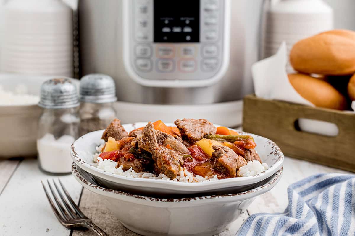 A serving of beef stew over rice in a white bowl with a pressure cooker in the background.