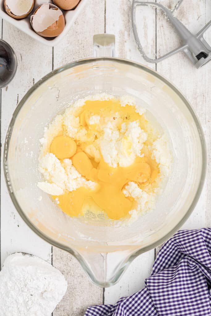 Glass bowl with eggs and sugar mixture, mixer beater, eggshells, and checked cloth on white surface.