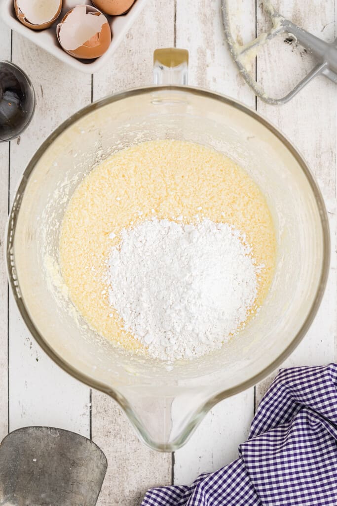 Large mixing bowl with flour on top of creamed butter and sugar, surrounded by eggshells and a blue checkered cloth.